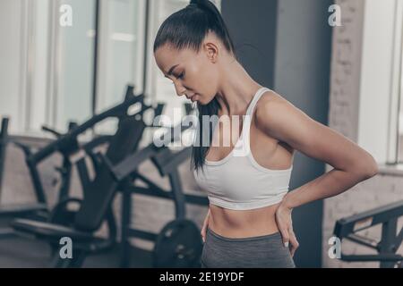 Schöne athletische Frau mit Rückenschmerzen nach dem Training im Fitnessstudio. Attraktive Fitness-Frau mit Rücken- oder Wirbelsäulenverletzung. Sportlerin, die sich den Rücken reibt Stockfoto