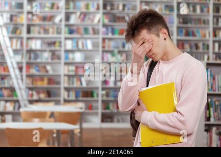 Junger müder Mann, der sein Gesicht reibt, an der Bibliothek steht und Platz kopiert. Erschöpft männlichen Schüler verlassen Bibliothek nach dem Studium für Stunden. Abschluss, z. B. Stockfoto