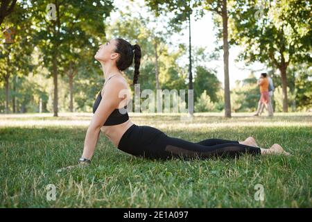 Athletische junge Frau, die im Park draußen trainiert, Cobra Asana tut Stockfoto