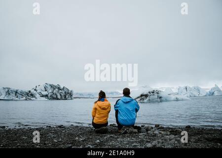 Nicht erkennbares Paar, das auf dem Rücken sitzt. Blick auf einen Gletscher in Island. Gelbe und blaue Mäntel. Konzept des Reisens und der Liebe Raum für Text. Stockfoto