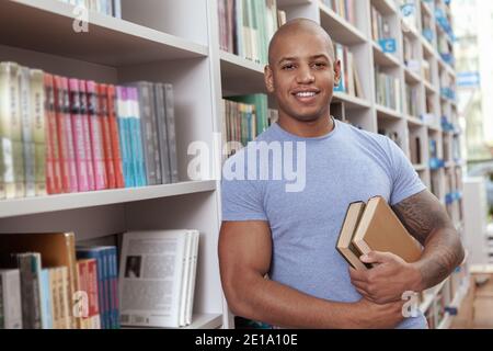 Fröhlicher, gutaussehender afrikanischer Mann, der zur Kamera lächelt, Bücher hält und sich an ein Bücherregal in der Bibliothek lehnt. Attraktiver junger Mann, der beim Buchmacher einkauft Stockfoto
