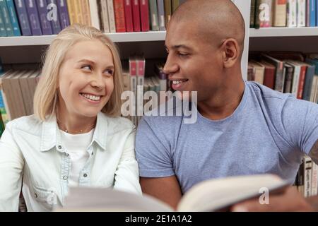 Nahaufnahme eines glücklichen jungen multiethnischen Paares, das jeden anlächelt Andere beim gemeinsamen Lesen eines Buches in der Bibliothek Stockfoto