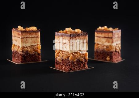 Vorderansicht von Stücken von frischem Dessert mit Biscuit und Mousse isoliert auf schwarzem Hintergrund geschichtet. Nahaufnahme von drei quadratischen Scheiben süßen braunen Schokoladenkuchen mit glasierten Top und Nüssen. Stockfoto