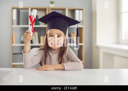 Nettes kleines Mädchen im akademischen Hut sitzend am Schreibtisch und Sie hält ihr Diplom Stockfoto