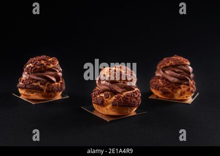 Drei leckere frische knusprige Profiterolen mit süßer brauner Schokoladencreme im Inneren. Nahaufnahme von hausgemachten leckeren gebackenen Eclirs isoliert auf schwarzem Hintergrund. Konzept der Desserts, Restaurant Essen. Stockfoto