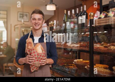 Glücklicher, gutaussehender junger Mann, der lachend in der Bäckerei arbeitet und frisches Brot verkauft Stockfoto