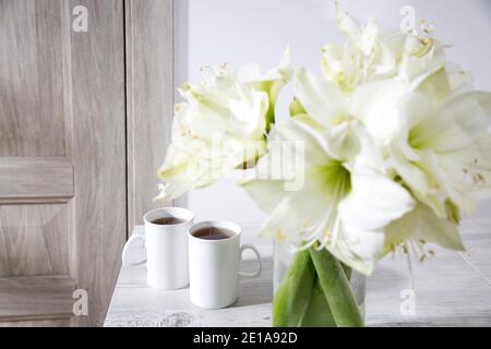 Bouquet von weißen Lilien in einer hohen Glasvase auf einem beigen Tisch an einer grauen Wand. Speicherplatz kopieren Stockfoto