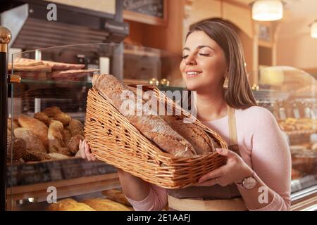 Schöne junge Unternehmerin, die gerne in ihrer Bäckerei arbeitet und köstliches frisch gebackenes Brot in einem Korb riecht Stockfoto