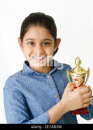 Portrait von einem schönen und lächelnden indischen Schulmädchen in weißem Hintergrund mit einer goldenen Trophäe und Darstellung Sieg. Stockfoto