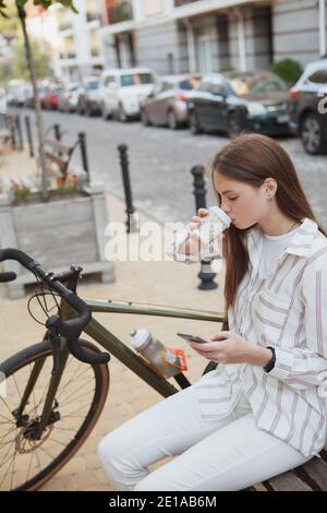 Vertikale Aufnahme einer jungen Geschäftsfrau, die Kaffee trinkt, Smartphone benutzt, auf einer Bank in der Nähe ihres Fahrrads in der Stadt sitzt Stockfoto