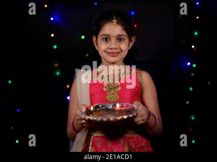 Ein schönes kleines indisches Mädchen in ethnischer Kleidung mit Juwelen gekleidet und hält Öllampen während diwali in der Nacht mit schwachen Lichtern im Hintergrund. Stockfoto