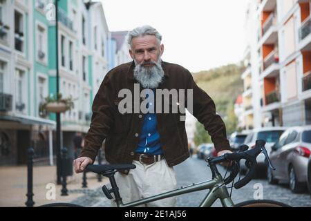 Gutaussehender älterer bärtiger Mann, der selbstbewusst auf die Kamera posiert Mit dem Fahrrad auf der Stadtstraße Stockfoto