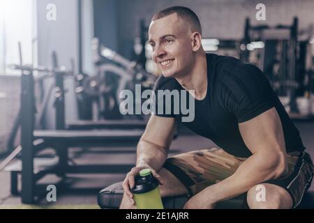 Glücklicher, gutaussehender junger Fitnessmann lächelnd, freudig wegblickend, sich nach dem Training im Fitnessstudio ausruhend. Fröhlicher Athletic Mann in Sportswear hält seinen shak Stockfoto