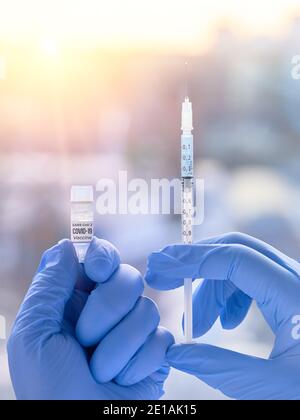 Impfstoff gegen Covid-19, verursacht durch das neuartige Coronavirus SARS-CoV-2. Hände in blauen Nitril-Handschuhen mit Spritze und Impffläschchen. Abends Blick über die Stadt Stockfoto