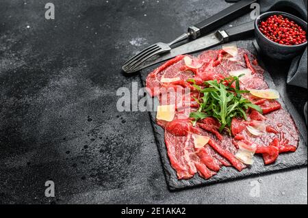 Rindercarpaccio auf schwarzem Teller mit Parmesankäse und Rucola. Schwarzer Hintergrund. Draufsicht. Speicherplatz kopieren. Stockfoto