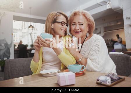 Fröhliche ältere Frau lächelt zur Kamera und umarmt ihre Erwachsene Tochter im Café Stockfoto