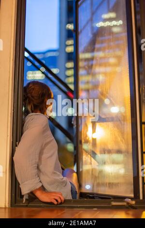 Die ästhetische Aufnahme zeigt eine junge Frau, die auf einer Fensterleiste sitzt, von hinten aufgenommen, während sie auf die Lichter der Stadt im alten Hafen von Montreal blickt. Stockfoto