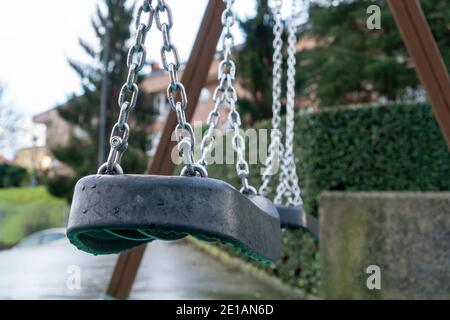 Leere Schwenkkette aus der Nähe mit Regentropfen auf einem Kunststoffsitz. Dramatische Aussicht. Ein Spielplatz in einem alten Stadtpark mit moosigen Betonwänden und Nadelbäumen Stockfoto