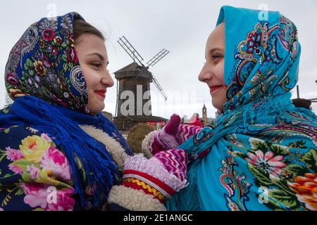 Susdal, Russland. 12. März 2016 EIN Mädchen hilft, einen Schal mit einem traditionellen russischen Muster auf dem Hintergrund einer Holzmühle in der Stadt Susdal in der Region Vladimir während festlicher Ereignisse, Russland zu binden. Susdal ist Teil der touristischen Route des Goldenen Rings Russlands, die durch die alten Städte Nordostrusslands führt, wo einzigartige Denkmäler der russischen Geschichte und Kultur erhalten sind Stockfoto