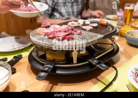 Fleisch Grillen und Käse Kochen in Pfannen auf einem elektrischen Raclette-Tischgrill in der Mitte eines Tisches Silvesterparty in Österreich Stockfoto
