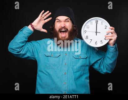 Ein gestresster junger bärtiger Mann hält eine große weiße Uhr und schaut sehr gestresst auf die Kamera in der Nähe einer schwarzen Wand. Stockfoto