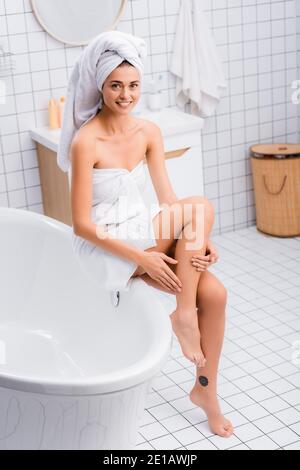 Glückliche Frau, eingewickelt in weiße Handtücher, Anwendung Körperlotion auf Bein, während auf der Badewanne sitzen Stockfoto