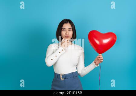 Nachdenklich Stirnrunzeln ernsthaft denkende asiatische Frau mit Hand unter dem Kinn, hält roten Herz-förmigen Ballon isoliert auf blauem Hintergrund Stockfoto