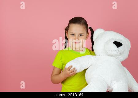 Nahaufnahme ein fröhliches, charmantes Mädchen mit Zöpfen in einem hellgrünen T-Shirt hält einen weißen Teddybären an der Pfote , isoliert auf einem rosa Hintergrund Stockfoto