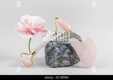 Crystal Rose Quarz Gesichtswalze und Massage-Tool Jade Gua Sha auf Steinen und rosa Blume auf grauem Hintergrund. Anti-Age-Gesichtsmassage für natürlichen Lift Stockfoto