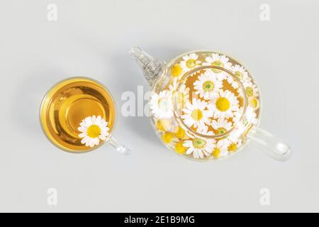 Tasse Tee und transparente Teekanne mit Kamillenblüten auf gelbem Hintergrund. Kamille Tee Vorteile Ihr Gesundheitskonzept. Draufsicht. Trendige Farben 2021 Stockfoto