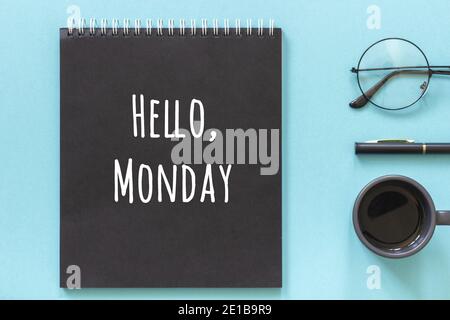 Hallo Montag Worte in schwarzem Notizblock, Stift, Tasse Kaffee und reeding Gläser auf blauem Hintergrund. Konzept Happy Monday. Draufsicht Flat Lay. Stockfoto