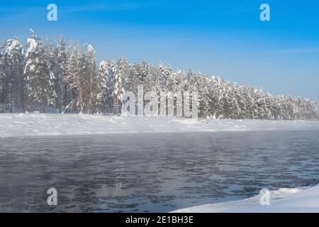 Frostiger Nebel über dem Winterfluss mit Schnee und Wald am Ufer. Erstes Eis am See an kalten Tagen. Stockfoto