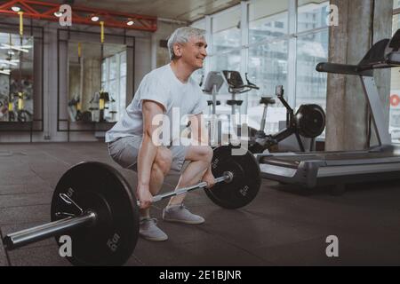 Ganzkörperaufnahme eines erfahrenen Sportlers, der mit Langhantel im Fitnessstudio arbeitet, Kopierbereich. Ältere Gewichtheber trainieren mit schwerer Langhantel. Gealterte Fitness m Stockfoto
