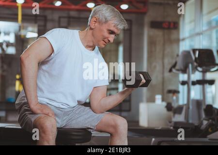 Starke gesunde ältere Mann tun Bizeps Übung mit einer Hantel, kopieren Raum. Glücklicher älterer Sportler, der im Fitnessstudio mit Gewichten arbeitet. Gesundheit, Acti Stockfoto