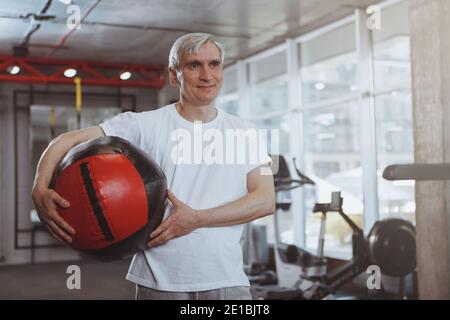 Glücklicher älterer, gesunder Mann, der freudig wegschaut, lächelt und den Medizinball hält. Entspannter, fröhlicher Sportler, der sich nach dem Training mit mir im Fitnessstudio ausruhte Stockfoto