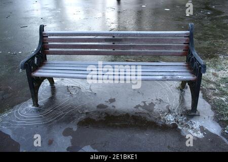 Nahaufnahme der verlassenen Parkbank mit Frost umgeben von gefrorenem Wasser und festem Eis um die Beine der Bank. Draußen im Winter Stockfoto