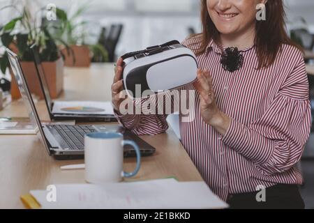 Eine zugeschnittene Aufnahme einer lächelnden Geschäftsfrau, die an ihrem Arbeitsplatz vor einem Laptop sitzt, eine 3D-Virtual-Reality-Brille in der Hand hält und Platz zum Kopieren bietet. Glückliche weibliche Entr Stockfoto