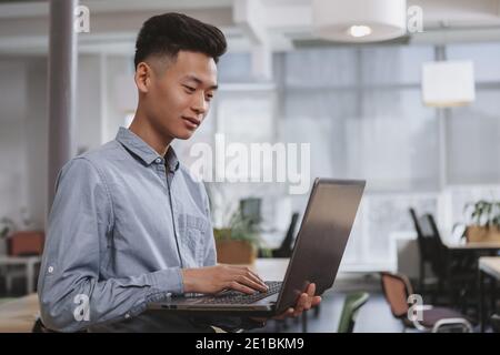 Junge asiatische männliche Student mit seinem Laptop, kopieren Raum. Gelegenheitskaufmann, der am Computer tippt und im Büro arbeitet. Männlicher Unternehmer, der daran arbeitet Stockfoto