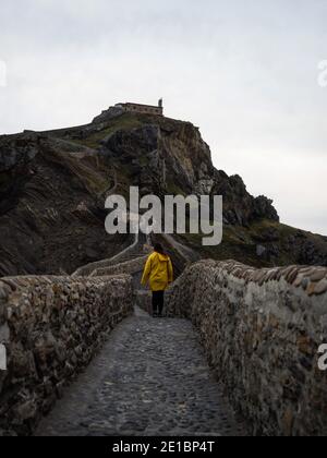 Frau in gelber Jacke stehend auf Steintreppe Fußgängerbrücke zu Gaztelugatxe Insel Felsenburg Einsiedelei Kapelle Insel Bermeo Baskenland Spanien Eu Stockfoto