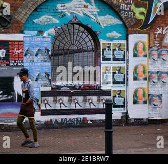 Mann mit Mütze und Schürze auf Spaziergängen vorbei an gegrilltem Bogenfenster in Ziegelwand mit Graffiti & Plakaten bedeckt, Shoreditch, East London, UK. Stockfoto