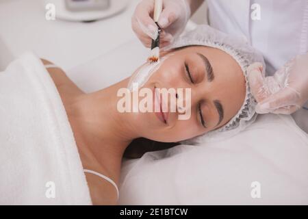 Gesunde glückliche Frau lächelt, während Kosmetologe Anwendung Gesichtsmaske auf ihre Haut. Schöne Cherful Frau genießen Gesichtsmaske Behandlung im Schönheitssalon Stockfoto