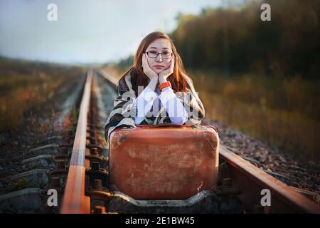 Eine schöne junge Frau in Brille und mit einem alten braunen Koffer sitzt auf den Gleisen und wartet leider auf die Ankunft des Zuges, um irgendwo hinzufahren. Jou Stockfoto