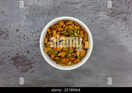 Draufsicht auf eine kleine weiße Schüssel, gefüllt mit Bio-Kürbiskernen und dehydrierten Apfelstückchen auf einer grau melierten Tischplatte. Stockfoto