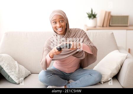 Fröhliche schwarze Dame in Hijab mit Controller spielen Videospiele auf Sofa zu Hause Stockfoto