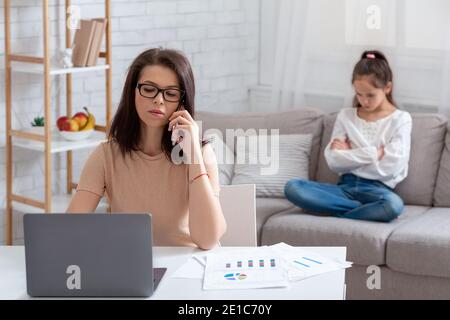 Junge Mutter arbeitet online, mit Laptop und Telefon, verärgert Kind sitzt auf dem Sofa in der Nähe, leiden unter Vernachlässigung Stockfoto