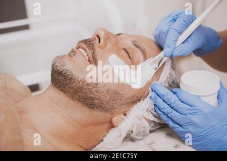 Reife bärtige freudigen Mann erhalten Gesichts-Spa-Behandlung, Kosmetikerin Anwendung Maske auf das Gesicht des männlichen Kunden. Glücklicher Mann, der sich im Spa Center entspannt, getti Stockfoto