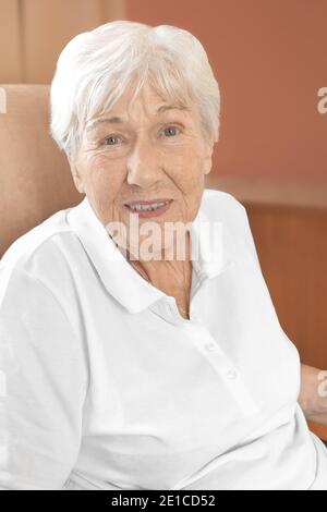 Fröhlich lächelnde ältere Frau in einem bequemen Stuhl in ihrem Zimmer in einem Altersheim. Stockfoto