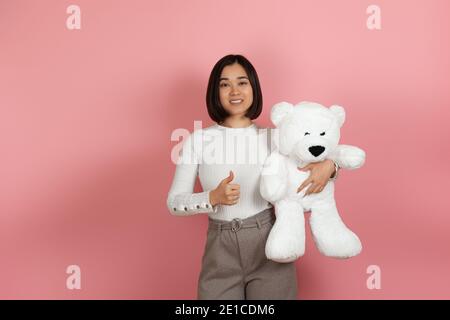 Lächelnd fröhlich asiatische Frau gibt einen Daumen nach oben und hält Ein weißer Teddybär isoliert auf einem rosa Hintergrund Stockfoto