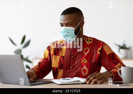 Arbeit Während Der Pandemie. Schwarz Freelancer Guy In Maske Mit Laptop Zu Hause Stockfoto