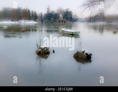 Schneebedecktes Boot im See und Love Temple im Hintergrund. Daumesnil-See (Wald von Vincennes, Paris, Frankreich). Weicher Fokus und gebleichte Winkel. Stockfoto